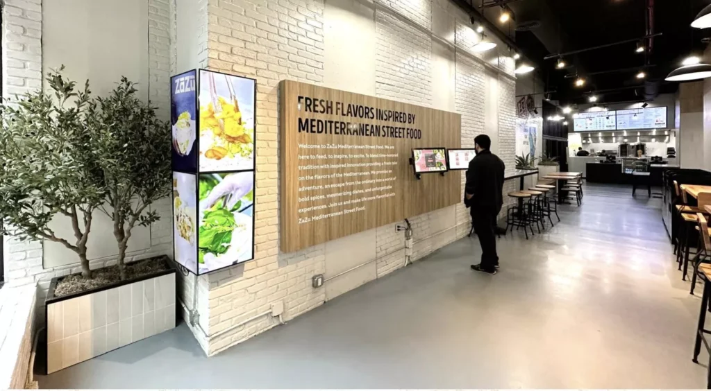 A long view of the interior wall at Zazu Mediterranean restaurant with a man reading the standee menu, and a plaque on the wall about Mediterranean Street Food.